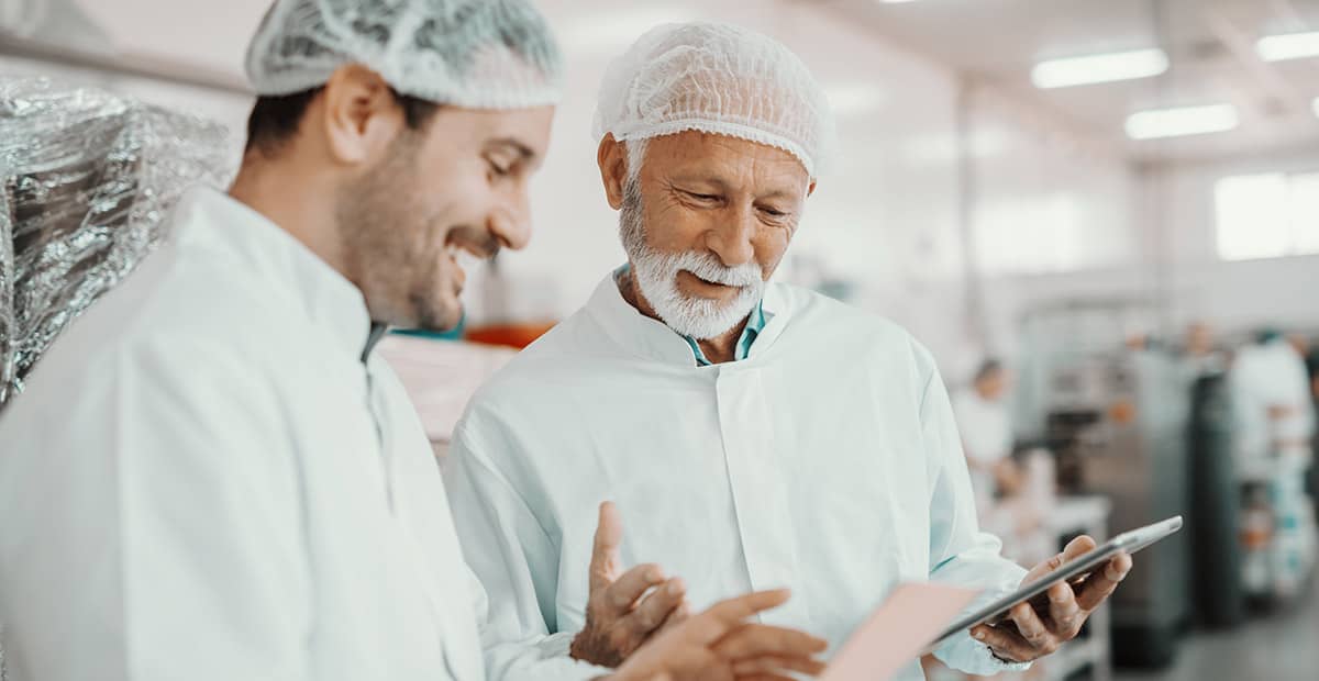 Two supervisors discussing about quality of food in food plant. Younger one holding folder with data while older one holding tablet. Both are dressed in white sterile uniforms and having hairnet.